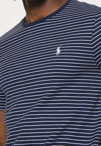 Námořnické pruhované tričko vyrobené z bavlny, s bílými vodorovnými pruhy, kulatým výstřihem a malým bílým logem na levé horní straně.