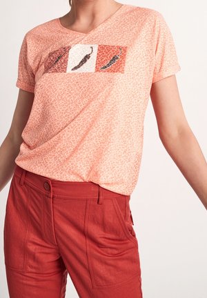 Nainen, joka käyttää persikanväristä leopardikuosista t-paitaa, jossa on chili-paprika-kuvioita, ja korkeavyötäröisiä ruosteenpunaisia housuja, seisoo yksinkertaisen taustan edessä.