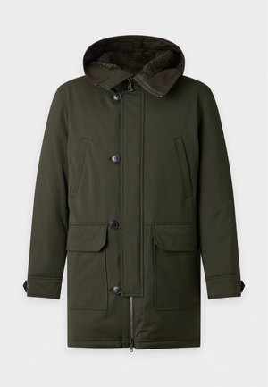 Un homme porte un manteau à capuche vert olive sur des vêtements noirs et des chaussures de randonnée bordeaux, devant un fond de studio uni.