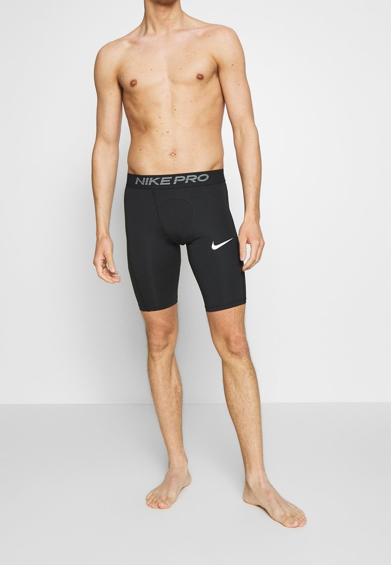 En shirtlös person som bär svarta Nike Pro kompressionsshorts med logotyp på vänster lår, stående barfota på ett ljust grått golv mot en vit vägg.