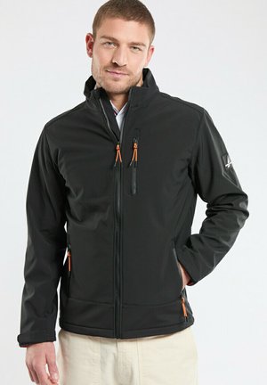 Homme portant une veste noire zippée avec des tirettes de couleur orange et un pantalon beige, debout avec une main dans la poche sur un fond uni.