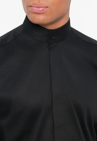 Černá košile s kravatu a slim fit, se zapínáním na knoflík u krku a hladkou, lesklou texturou látky.