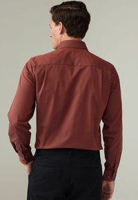 Chemise à manches longues bordeaux avec un motif texturé, col ouvert et poignets à boutons. Associée à un pantalon noir, vue de dos.