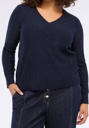 Femme portant un pull bleu marine à col en V moucheté et un pantalon en jean bleu foncé taille haute avec boutons, mains dans les poches.