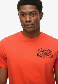 Camiseta de algodón roja con cuello redondo, mangas cortas y un diseño gráfico en la parte delantera con montañas y el texto "Superdry" en azul marino.