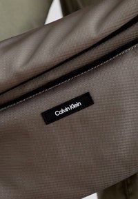 Calvin Klein szürke anyagú deréktáska, textúrázott felülettel, cipzáras záródással és fekete logós párnával az elülső részén.
