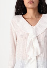 Blusa blanca con textura, cuello con volantes y botones, que presenta un diseño en capas y un material ligero, ligeramente transparente.