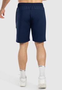 Marineblauwe sportshorts met een elastische tailleband en zijzak, vervaardigd van een gladde stof en met een relaxte pasvorm, gedragen met witte sneakers.