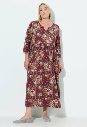 Brombeerfarbene floral bedruckte Maxikleid aus weichem Stoff mit Dreiviertelärmeln, das farbenfrohe Blumenmuster und eine lässige Silhouette aufweist. Kombiniert mit hellblauen Flats.