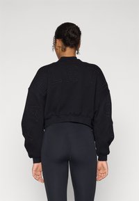 Crni skraćeni dukserica s vezom "Fabletics" na rukavima, okruglog izreza i rebrastim rubovima, u kombinaciji s crnim tajicama.
