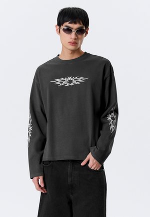 Mladý muž v čiernej dlhorukávovej košeli s bielymi plameňovými vzormi na hrudi a rukávoch, čiernych nohaviciach a striebornej slnečných okuliaroch.