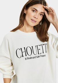 Krém színű pulóver, fekete hímzett felirattal, amelyen az szerepel: "CHOUETTE 86 Boulevard Saint Tropez". Lazább fazonú, eséses vállakkal.