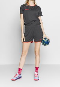 Conjunto deportivo gris con detalles rojos, que incluye una camiseta de manga corta y pantalones cortos. La modelo sostiene un balón de balonmano azul y verde, y lleva calzado colorido.