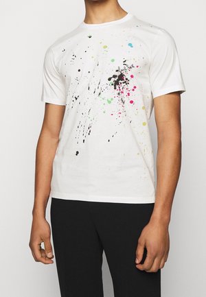 Mann trägt ein weißes T-Shirt mit kurzen Ärmeln und buntem Farbspritz-Design sowie schwarze Hose, steht vor einem einfarbigen Hintergrund.