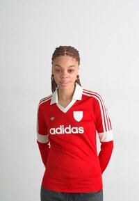Rød Adidas-drakt med hvite detaljer, tre striper på ermene og v-hals. Har en logo og et skjolddesign på brystet.