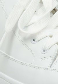 Witte sneaker met een gladleren bovenwerk, ronde veters en subtiele stikseldetails. De oogjes zijn grijs; de veters zijn plat en textuur.