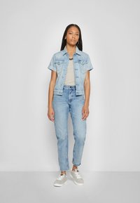 Chaleco de mezclilla azul claro con mangas cortas, llevado sobre una camiseta a rayas grises y blancas; combinado con jeans azul claro y zapatillas grises.