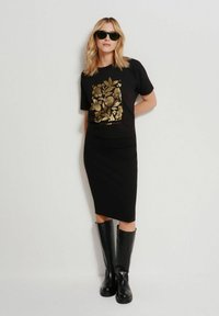 Czarna koszulka z złotym kwiecistym wzorem, w połączeniu z czarną ołówkową spódnicą i czarnymi botkami do kolan. Modelka nosi okulary przeciwsłoneczne.