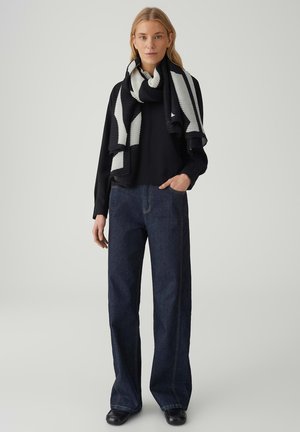 Haut noir à manches longues associé à un jean denim foncé à jambes larges. Une écharpe rayée noire et blanche en maille côtelée est drapée autour du cou. Chaussures noires.