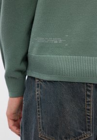 Grüner gestrickter Pullover mit strukturiertem Stoff, gerippten Bündchen und Saum. Auf der unteren Rückseite befindet sich bedruckter Text. Kombiniert mit dunklen Jeans.