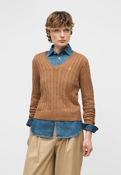 Νεαρό άτομο που φοράει καφέ πουλόβερ με πλεκτό σχέδιο πάνω από μπλε πουκάμισο με κουμπιά και μπεζ παντελόνι φαρδύ, στέκεται μπροστά σε απλό φόντο.