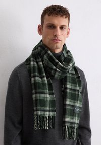 Écharpe à carreaux verts et noirs en tissu doux avec des bords frangés ; présente un motif texturé et une petite étiquette de marque d'un côté.