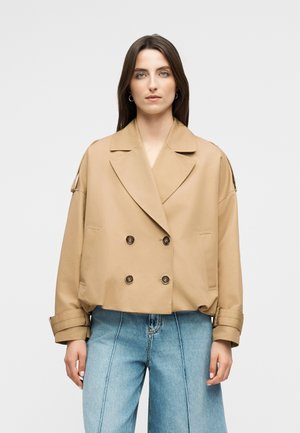 Γυναίκα με μπεζ κοντό σακάκι διπλής σειράς κουμπιών με φαρδιούς πέτου και ανοιχτό μπλε τζιν παντελόνι ψηλής μέσης, σε απλό φόντο.