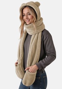 Beige fleece tørklæde med h�ætte, der har bjørneører, sidelommer og en blød, tekstureret overflade. Båret over en grå sweater.