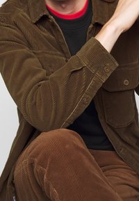 Camisa de pana marrón con grandes bolsillos, puños abotonados y acanalado vertical, llevada sobre una sudadera negra y pantalones de pana marrón.