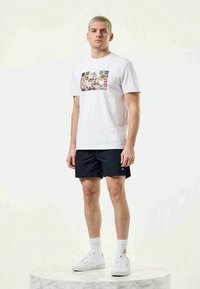 White cotton t-shirt with a graphic print featuring various logos, paired with black shorts, white sneakers, and white socks.
