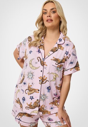Lyshåret kvinde iført pastelrosa kortærmet pyjamas s mønstre af tiger, måne, sol, stjerne og øje, står mod hvid baggrund.