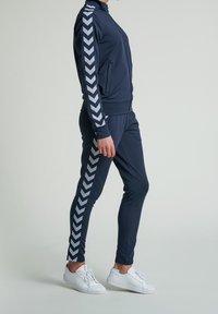 Laineen takki tummansinisellä värillä, jossa on valkoinen chevron-kuvio hihojen ja sivujen varrella. Takissa on täysipitkä vetskari, sivutaskut ja siihen sopivat kapeat housut.