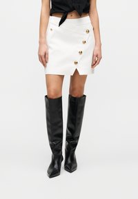 Λευκή μίνι φούστα τύπου wrap με χρυσά κουμπιά στο μπροστινό μέρος. Συνδυασμένη με μαύρες δερμάτινες μπότες μέχρι το γόνατο με μυτερά δάχτυλα.
