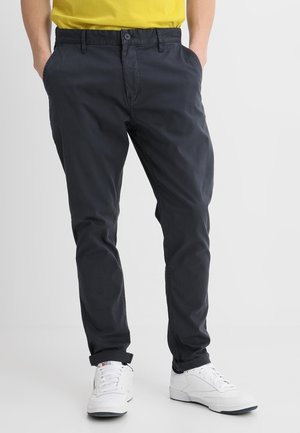 Homme portant un pantalon chino noir aux ourlets retroussés, des baskets blanches et une chemise jaune, les mains dans les poches, sur fond blanc.