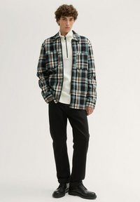 Karierte Flanellbluse in Schwarz, Weiß und Türkis mit zwei Brusttaschen, über einem cremfarbenen Pullover getragen, kombiniert mit schwarzen Hosen und Schuhen.