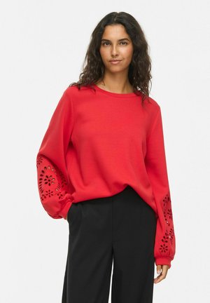 Femme aux longs cheveux bouclés portant un haut rouge à manches longues avec des découpes florales sur les manches et un pantalon noir, posant devant un fond uni.