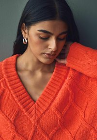 Helle orange gestrickte Sweater mit tiefem V-Ausschnitt, geribbeltem Kragen und strukturierten Diamantmustern. Modell trägt metallene Creolen.