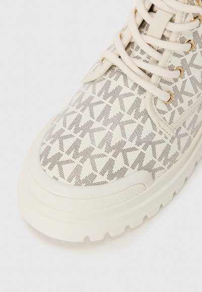 Vit sneaker med ett texturerat tyg som har ett upprepande mönstrat logo. Rundad tå, bruna snören och förhöjd gummisula med grepp.