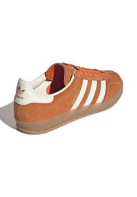 Suedesneaker i orange med hvidt tre-stribes design, gummisål og branding på hæl og tunge. Har polstret krave og snørebånd.