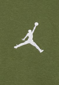 Un tessuto verde oliva mostra un'importante silhouette bianca ricamata di una figura che salta, mentre tiene un pallone da basket. La texture è liscia.