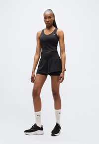 Αθλητική γυναίκα όρθια, φορώντας μαύρο αμάνικο τοπ, μαύρα στρωμένα σορτς, λευκές κάλτσες έως τον αστράγαλο και μαύρα παπούτσια για τρέξιμο, πάνω σε απλό ανοιχτό φόντο.