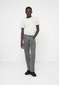 Jean gris à coupe droite, porté avec un t-shirt blanc orné d'un petit logo. Des chaussures noires complètent la tenue. Fond uni.