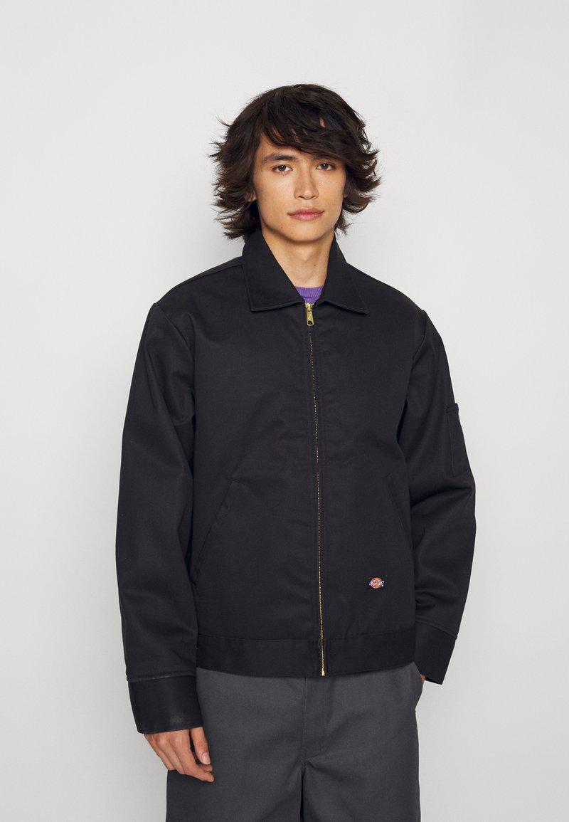 Chaqueta negra con cremallera, cuello tipo camisa, corte recto y puños elásticos. Presenta una cremallera dorada y un pequeño logotipo en el pecho.