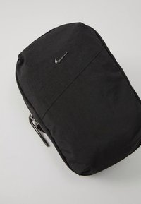 Bolsa preta da Nike com fecho de correr, textura de tecido e pequeno logótipo prateado da swoosh na frente, colocada plana numa superfície clara.