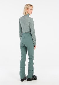 Maglietta verde a maniche lunghe e pantaloni da sci verdi abbinati con caviglie con zip, realizzati in un materiale liscio e elastico. Footwear è rappresentato da scarponi da sci neri.