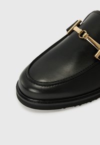 Chaussure en cuir noir avec une finition lisse, bout arrondi et un détail de boucle ton or sur la partie supérieure. Semelle en caoutchouc avec un motif de traction minimal.