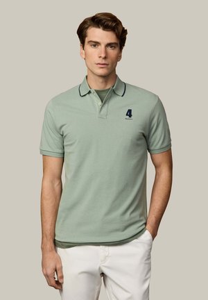 Jeune homme portant un polo vert clair avec le numéro 4 et une bordure noire sur le col, associé à un pantalon blanc, debout devant un fond uni.