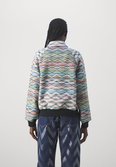 Pull-over coloré matelassé avec un motif en vagues bleu, vert, orange et blanc ; présente un ourlet et un col côtelés noirs. Associé à un jean texturé.