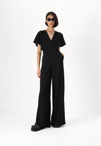Svart jumpsuit med korte fladdermer, dyp V-hals og en tettsittende midje. Buksebeina er utsvingte og når gulvet, kombinert med kraftige svarte platå sko.