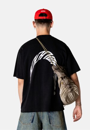 Mand iført en sort grafisk T-shirt, blå jeans, rød kasket og bærende på en beige snørelukningspose med et mønstret skulderbånd, set bagfra.